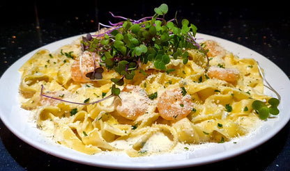 A close-up shot of a creamy pasta dish on a white plate, featuring shrimp, microgreens, and grated cheese on a dark background.